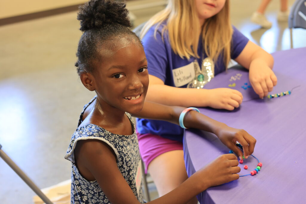 A young girl assembles a bracelet in the SMART Girls program while smiling at the camera