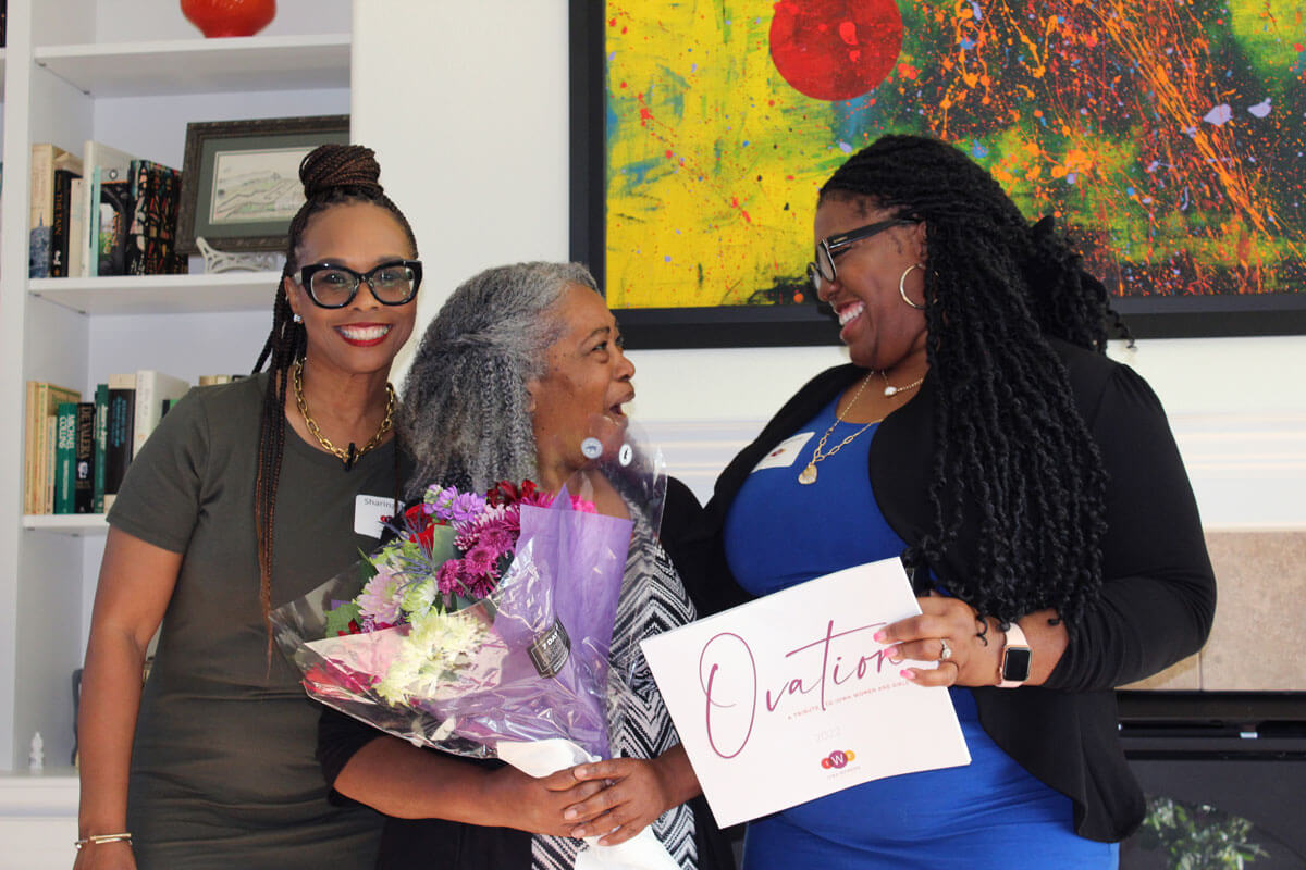 An older woman holding flowers is being presented a copy of Ovation, looking at the woman presenting it to her in a way of pleasant surprise